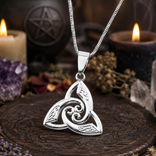 Silver Celtic triquetra trinity knot pendant necklace on curb chain, styled on dark carved wooden altar with pentagram, lit candles, amethyst crystal, and dried flowers