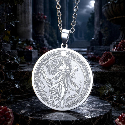 A silver pendant necklace featuring an intricately engraved fantasy illustration of a flowing gown and decorative border, hanging from a silver chain against a dark, atmospheric gothic stone background with pomegranate accents.