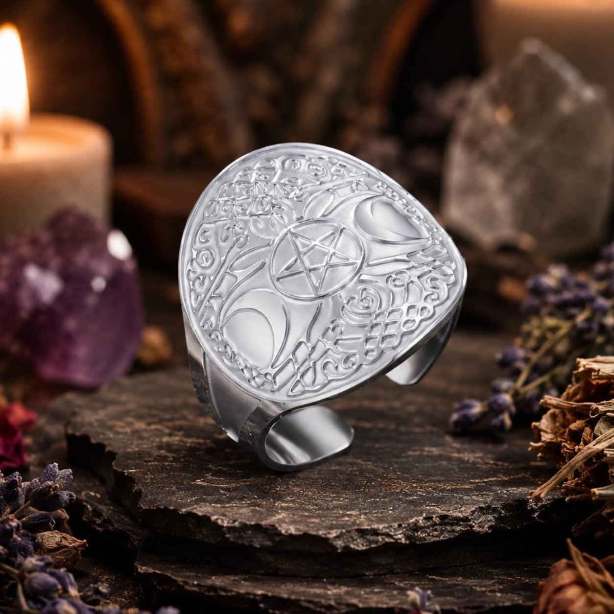 A silver-toned ring with an engraved pentagram and moon phases design, displayed on a dark wooden surface surrounded by candles, crystals, and dried lavender.