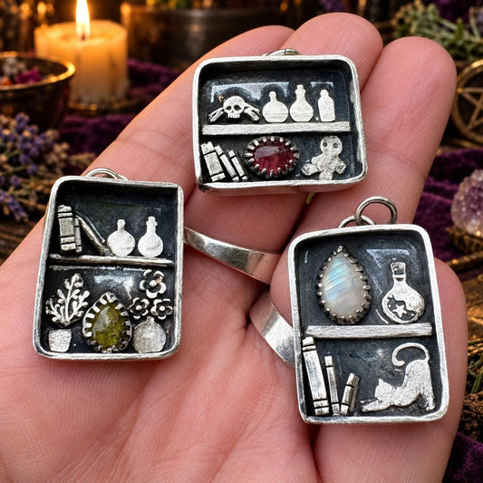 Two apothecary shelf pendant necklaces with potion bottles, books, cat and moonstone held in hand