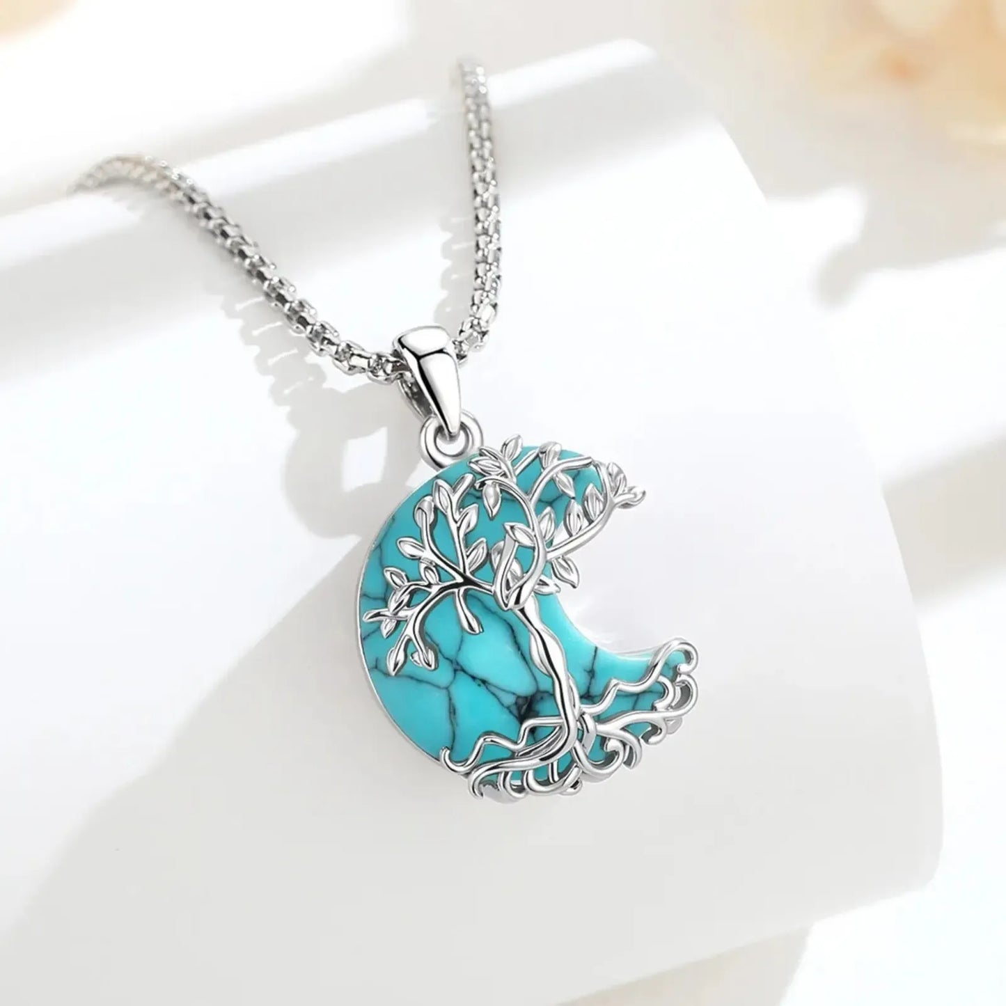 Turquoise Tree of Life crescent moon necklace displayed on white surface with soft floral background