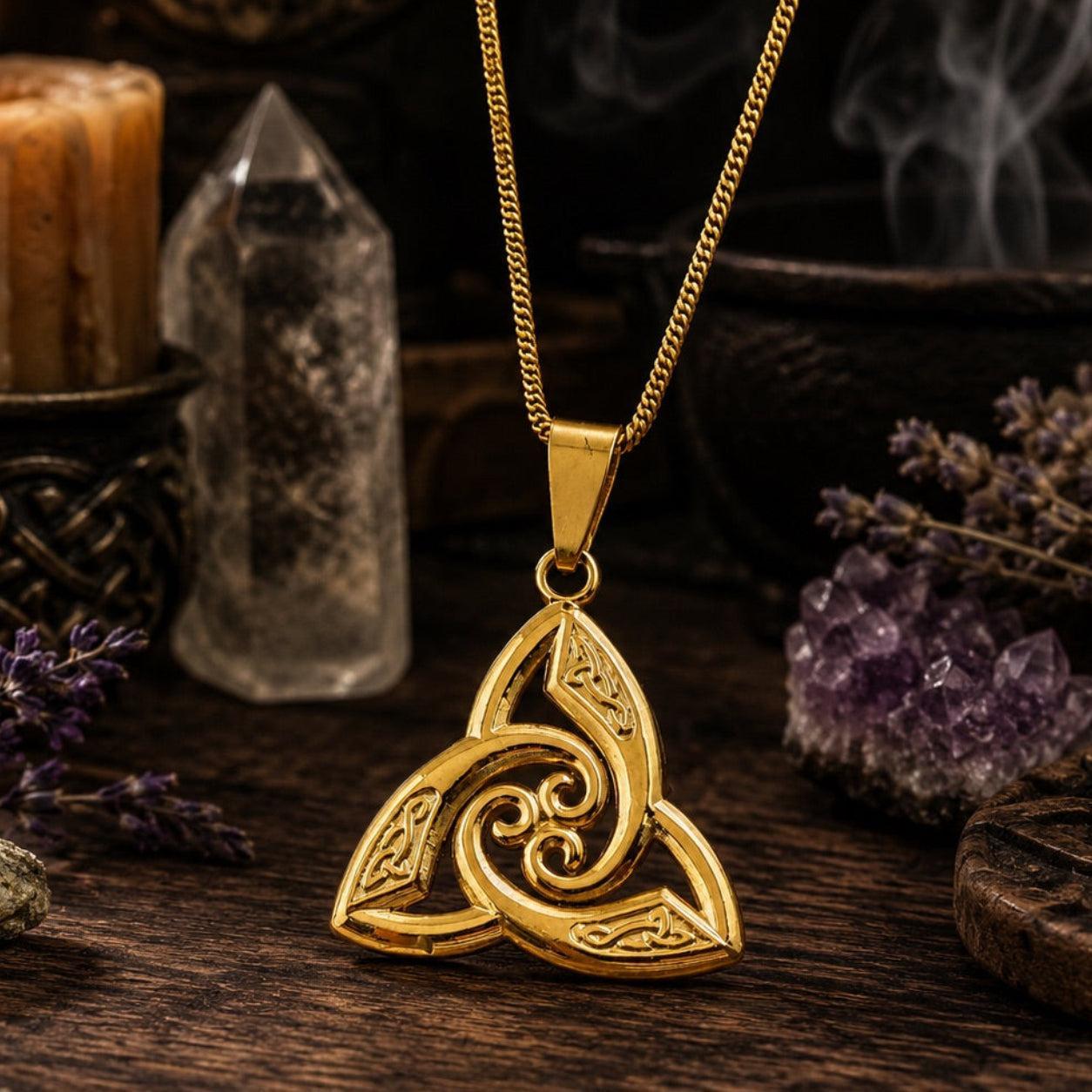 Gold Celtic triquetra trinity knot pendant necklace on cable chain, displayed on rustic wooden surface with quartz crystal point, amethyst cluster, lit candle, and dried lavender