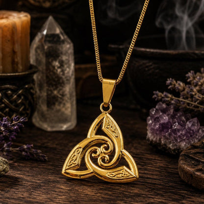 Gold Celtic triquetra trinity knot pendant necklace on cable chain, displayed on rustic wooden surface with quartz crystal point, amethyst cluster, lit candle, and dried lavender