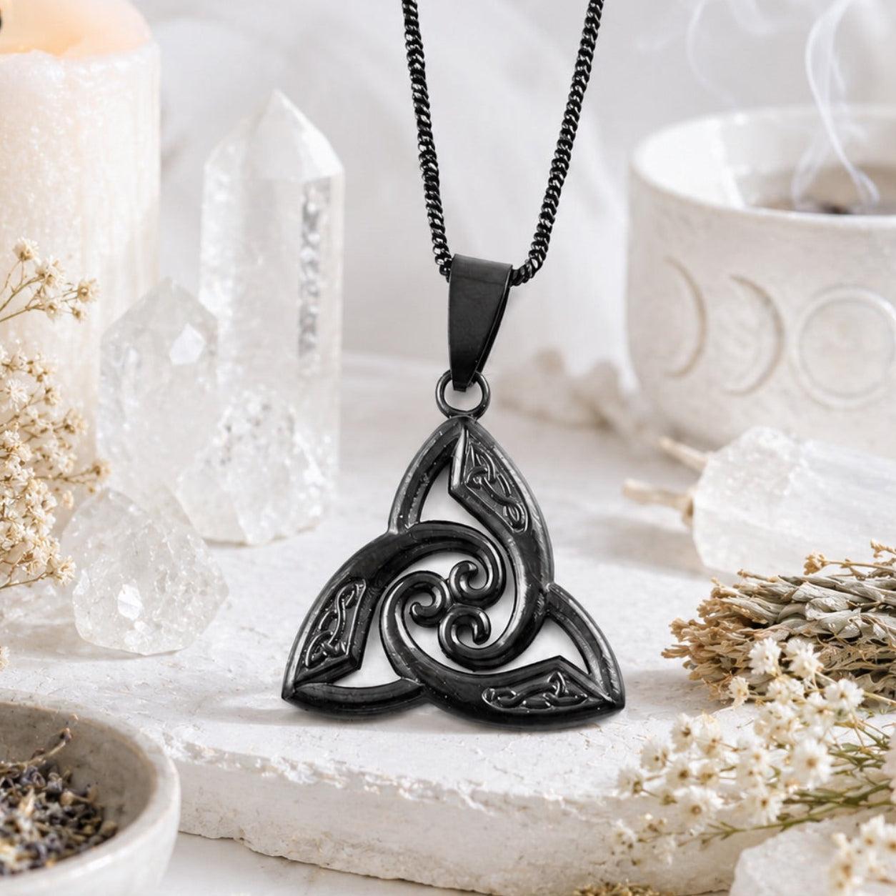 Black stainless steel Celtic triquetra trinity knot pendant necklace with spiral swirl design, displayed on white altar stone with quartz crystals and dried flowers