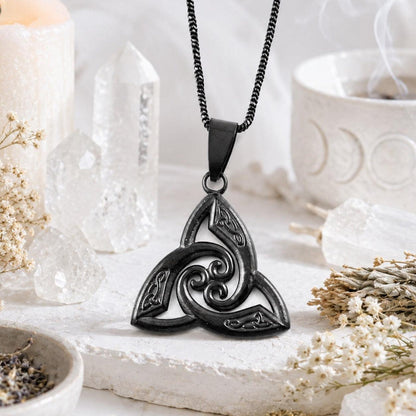 Black stainless steel Celtic triquetra trinity knot pendant necklace with spiral swirl design, displayed on white altar stone with quartz crystals and dried flowers