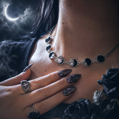 A gothic-style silver necklace with oval moon and planet charms worn on a model's neck, paired with matching gothic rings and long dark manicured nails, set against a dark moody background with a crescent moon.