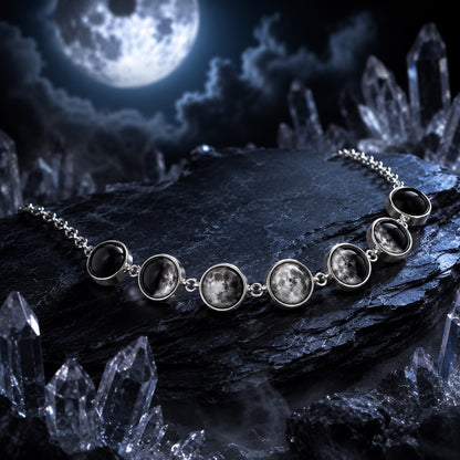 A silver-toned chain necklace featuring seven oval moon face pendants with detailed lunar imagery, displayed on a dark rock surrounded by crystal formations under a full moon sky.