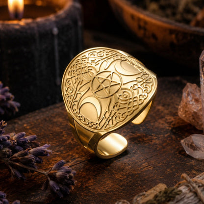 A gold-toned metal ring with an engraved shield design featuring a pentagram and moon phases, styled on a dark wooden surface with a lit candle, crystals, and dried lavender.
