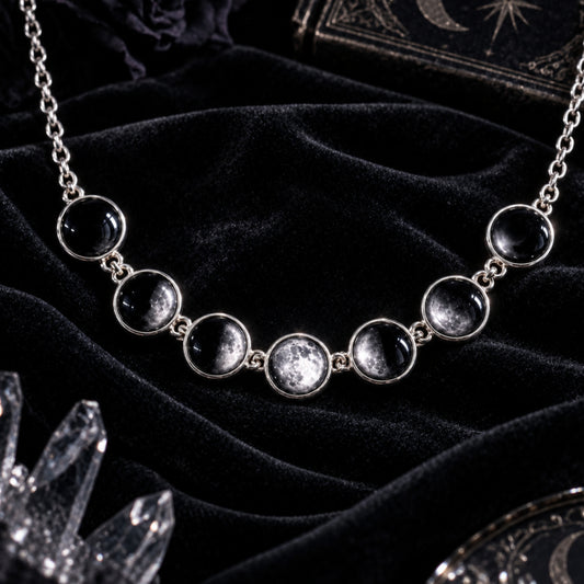 Celestial Moon Phase Choker necklace featuring seven black moon-shaped beads with silver settings and chain, displayed on a pedestal against a dark starry sky and crystal background.