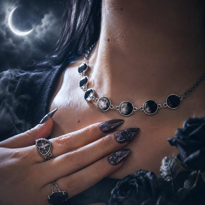 A gothic-style silver necklace with oval moon and planet charms worn on a model's neck, paired with matching gothic rings and long dark manicured nails, set against a dark moody background with a crescent moon.