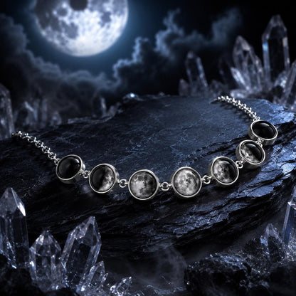 A silver-toned chain necklace featuring seven oval moon face pendants with detailed lunar imagery, displayed on a dark rock surrounded by crystal formations under a full moon sky.