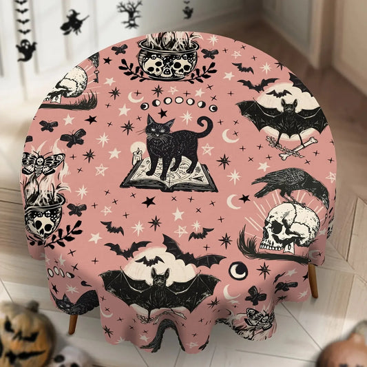 A round table covered with a pink Halloween-themed tablecloth featuring black illustrations of a cat on an open book, skulls, bats, ravens, stars, moons, and butterflies, shown in a styled indoor setting.