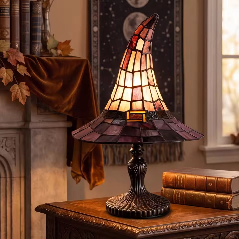 A stained-glass table lamp shaped like a witch's hat, glowing with warm amber and orange light, set on a wooden table in a cozy, warmly lit room next to a window and books.