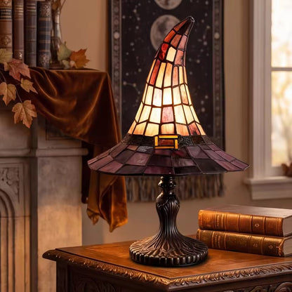 A stained-glass table lamp shaped like a witch's hat, glowing with warm amber and orange light, set on a wooden table in a cozy, warmly lit room next to a window and books.