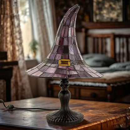 A stained glass table lamp shaped like a witch's hat, featuring purple and pink glass panels with a small gold accent band, set on a dark metal pedestal base on a wooden table in a cozy room.