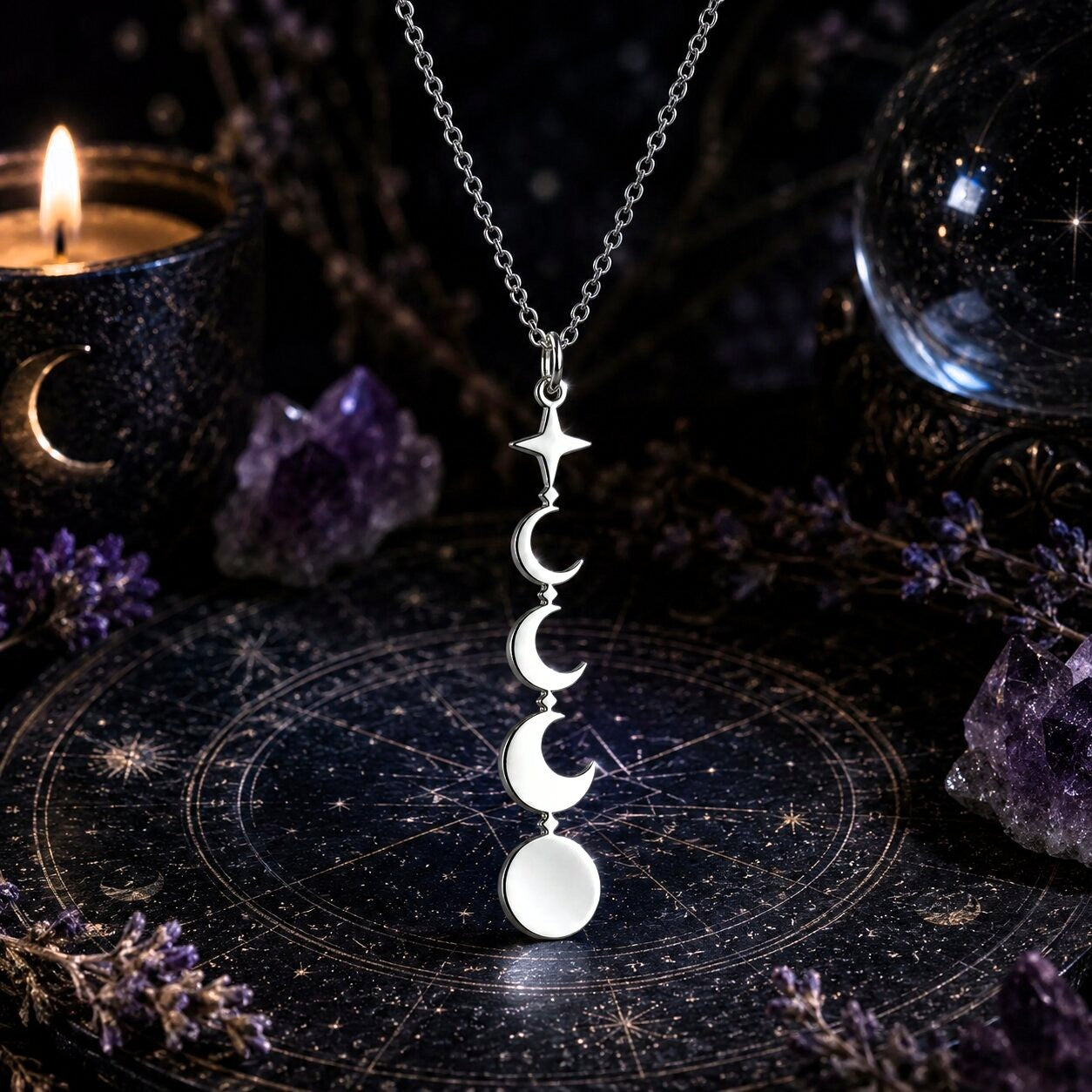 A silver necklace with a vertical triple-moon-and-star pendant is displayed on a dark celestial mat, surrounded by purple crystals, dried flowers, a lit candle, and a glass orb.