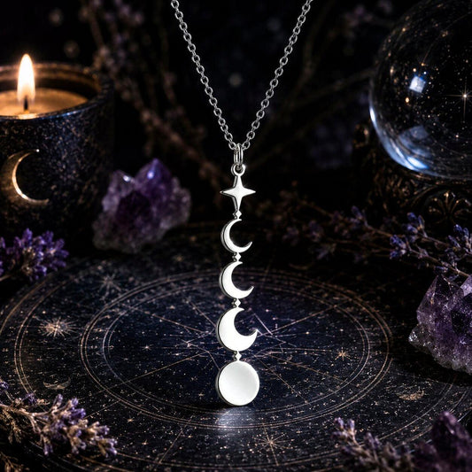 A silver necklace with a vertical triple-moon-and-star pendant is displayed on a dark celestial mat, surrounded by purple crystals, dried flowers, a lit candle, and a glass orb.