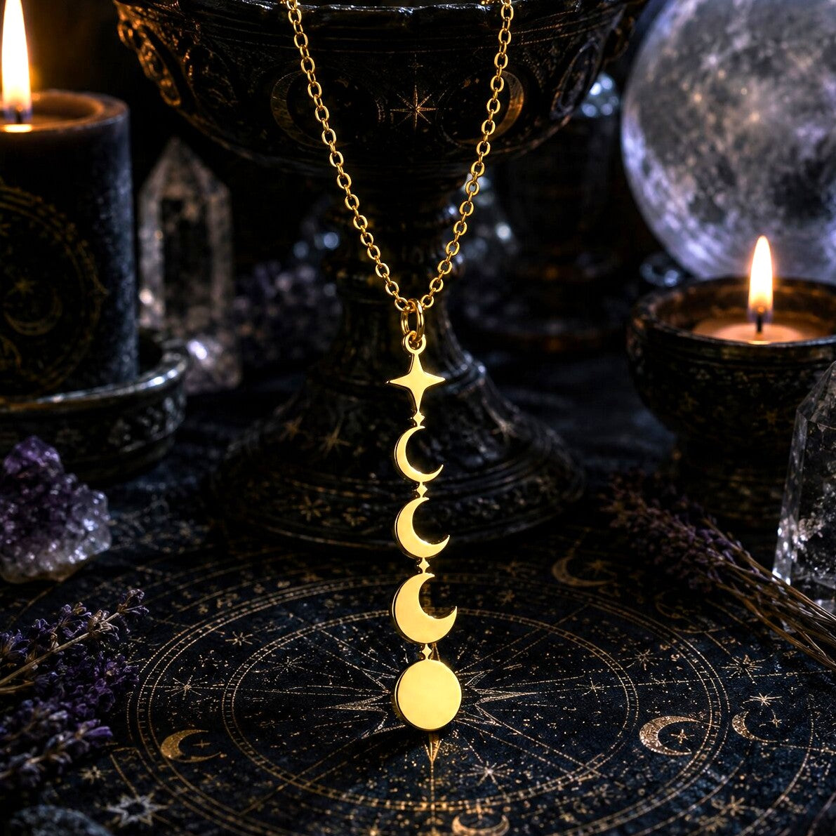 A gold necklace with a vertical triple-moon and full moon pendant is displayed on a dark celestial mat, surrounded by candles, crystals, and a black pedestal bowl.