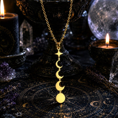 A gold necklace with a vertical triple-moon and full moon pendant is displayed on a dark celestial mat, surrounded by candles, crystals, and a black pedestal bowl.