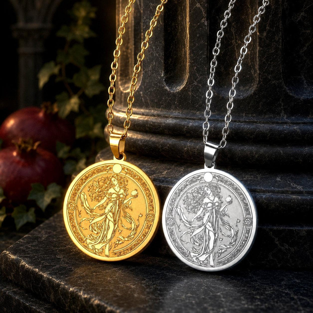 Two coin-style pendant necklaces with an engraved female figure, one in gold and one in silver, displayed on a dark stone pedestal with pomegranate props in the background.