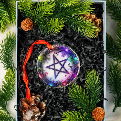 Clear glass pentagram ornament with colorful lights and a red ribbon, nestled in a gift box with pine branches.