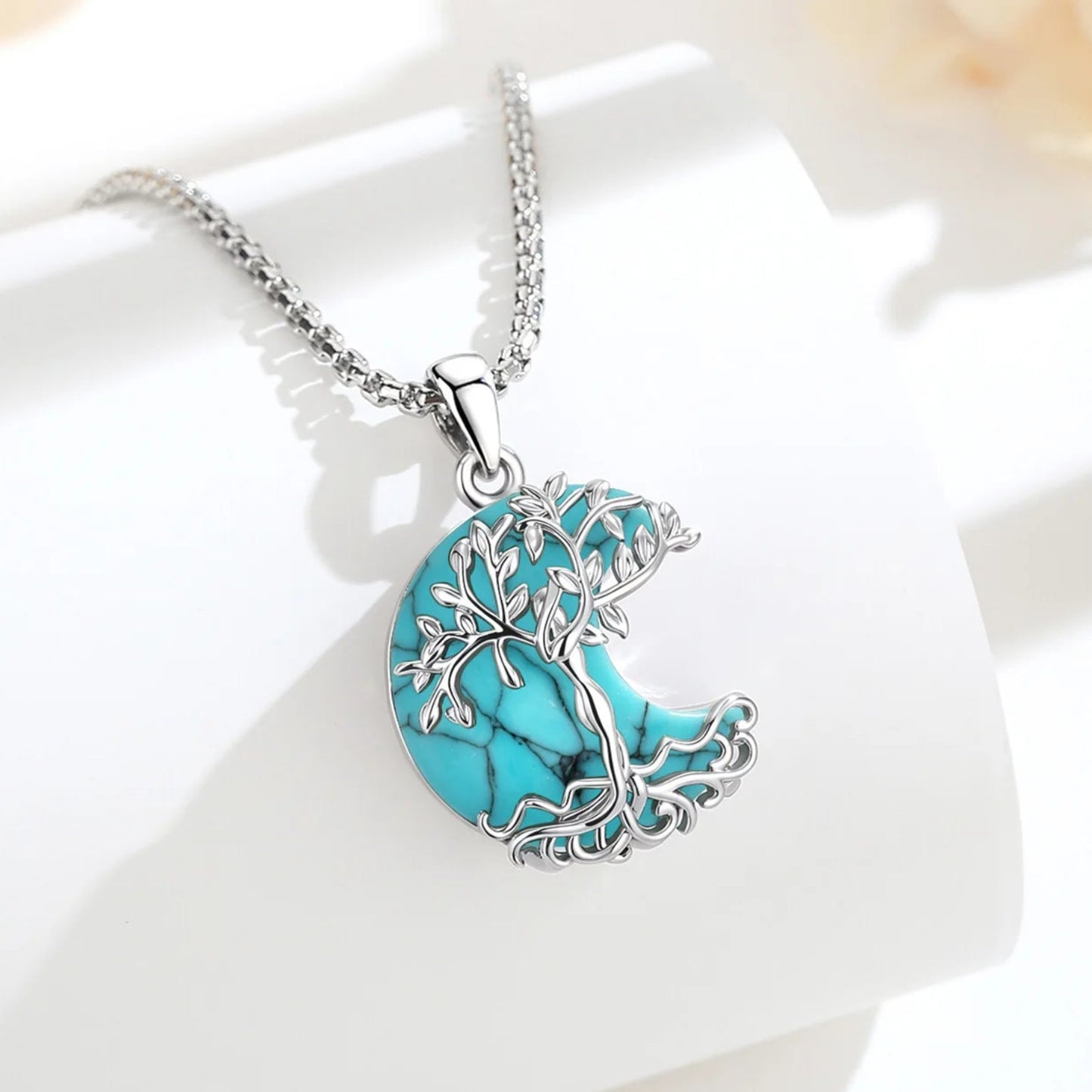 Turquoise Tree of Life crescent moon necklace displayed on white surface with soft floral background