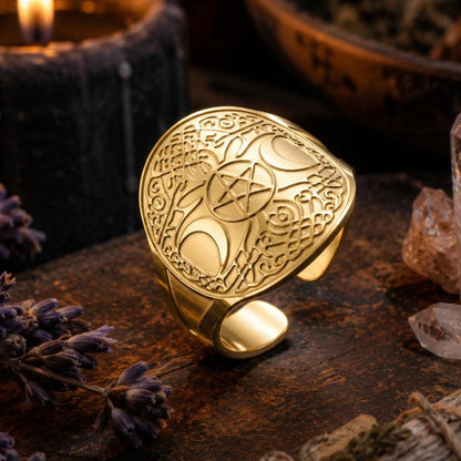 A gold-toned metal ring with an engraved shield design featuring a pentagram and moon phases, styled on a dark wooden surface with a lit candle, crystals, and dried lavender.