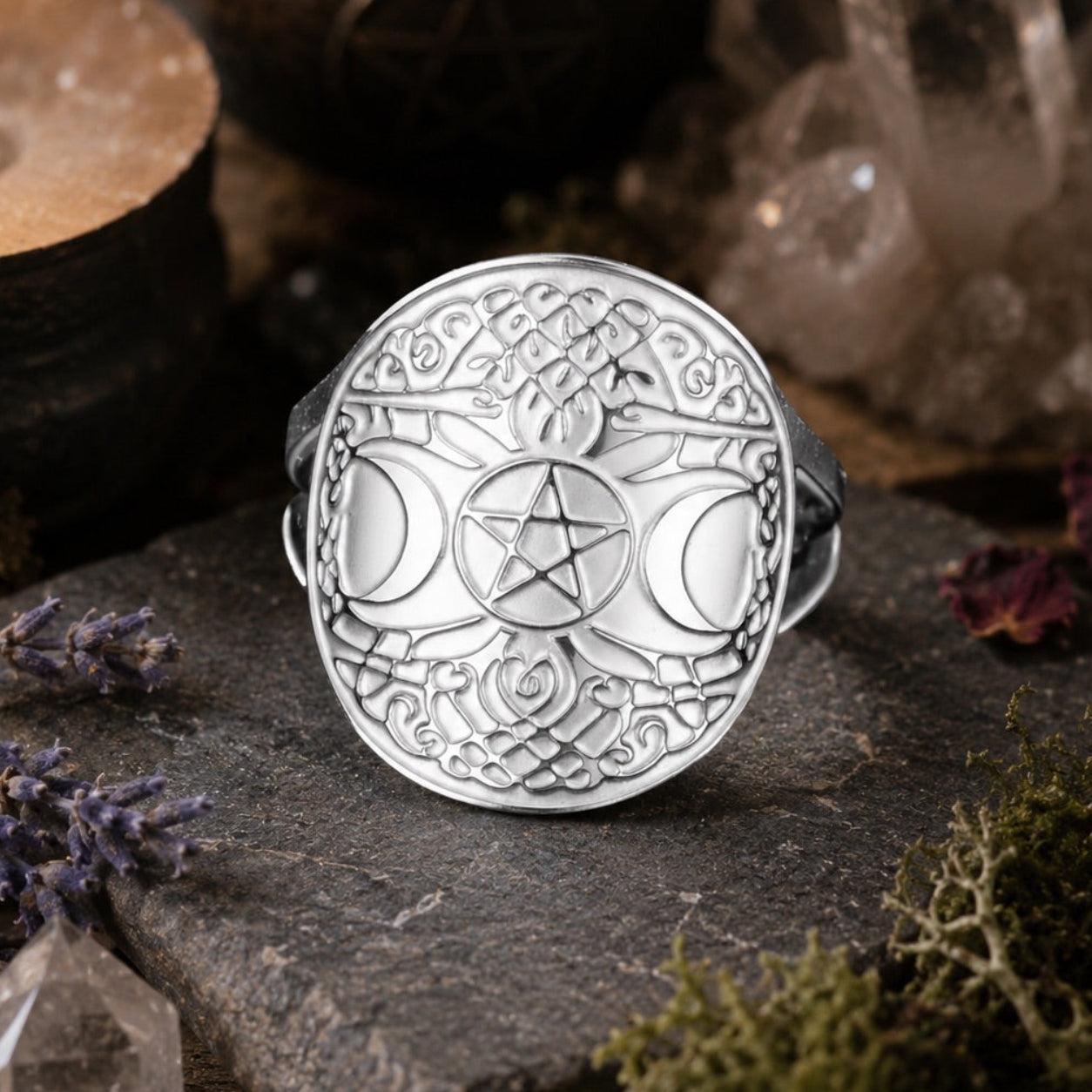 A silver-toned triple moon and pentagram ring displayed on a dark stone slab, surrounded by mystical props including a burning candle, crystals, dried flowers, and moss.