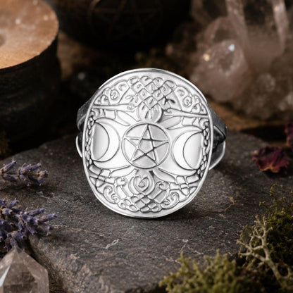 A silver-toned triple moon and pentagram ring displayed on a dark stone slab, surrounded by mystical props including a burning candle, crystals, dried flowers, and moss.