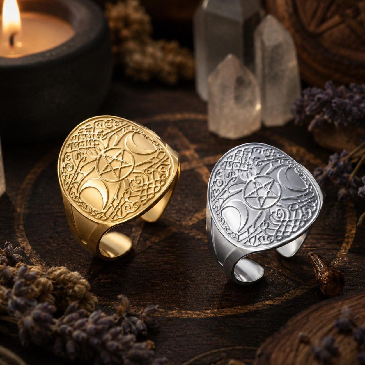 Two engraved statement rings displayed on a dark wooden surface with mystical props. One ring is gold-toned and the other is silver-toned, both featuring a large crescent moon and pentagram design.