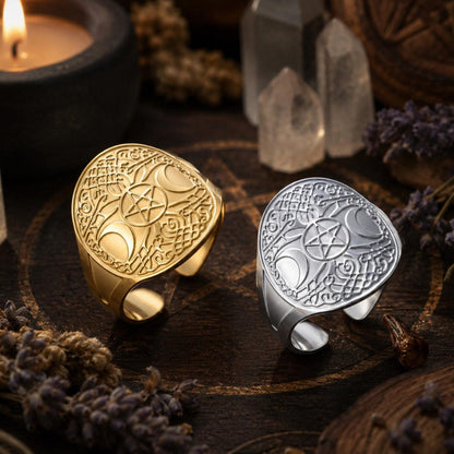 Two engraved statement rings displayed on a dark wooden surface with mystical props. One ring is gold-toned and the other is silver-toned, both featuring a large crescent moon and pentagram design.