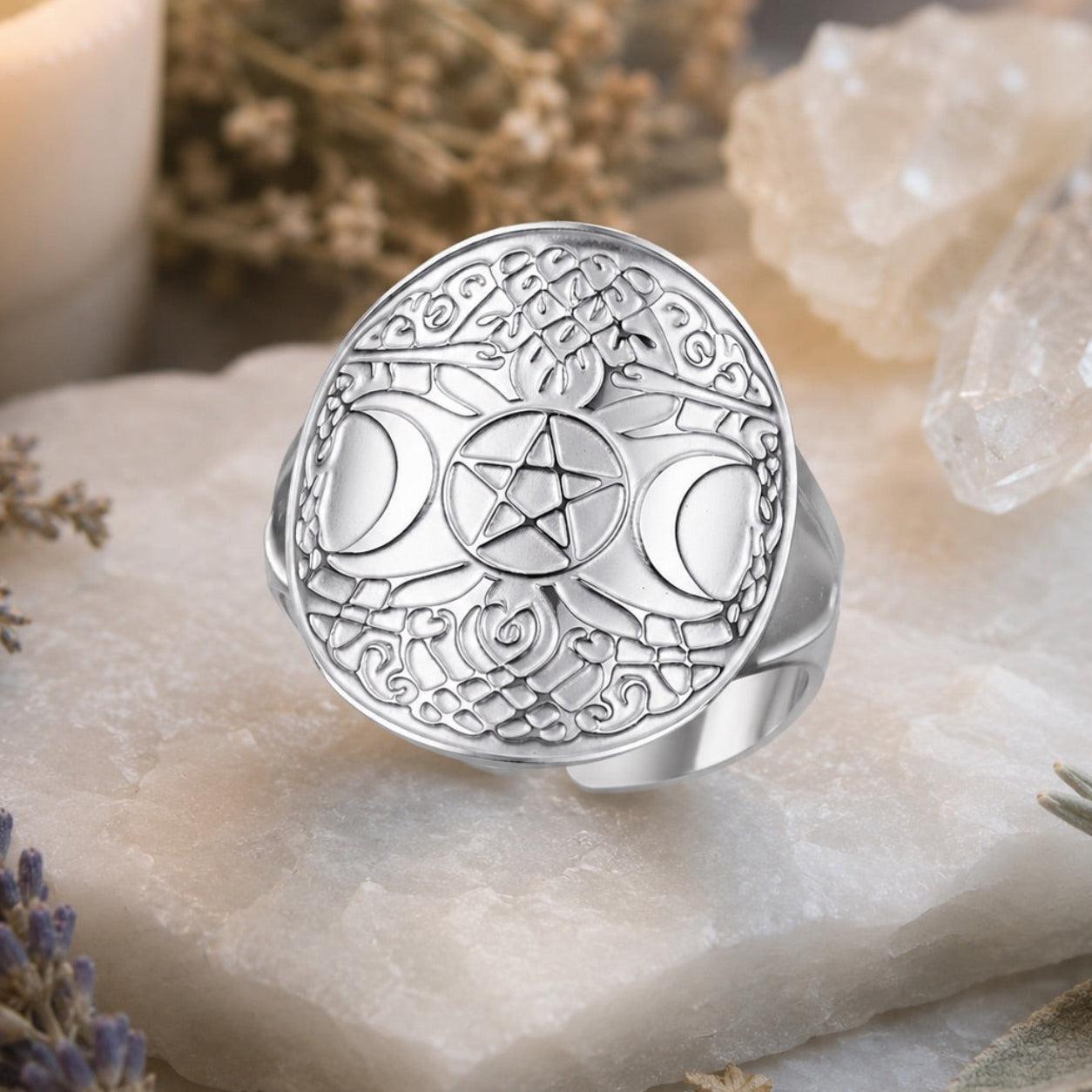 A silver-toned ring with an intricate engraved celestial design, featuring a central pentagram and moon phases, displayed on a white stone slab with dried flowers and crystals.