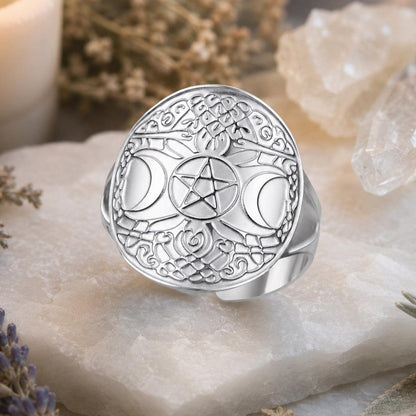 A silver-toned ring with an intricate engraved celestial design, featuring a central pentagram and moon phases, displayed on a white stone slab with dried flowers and crystals.