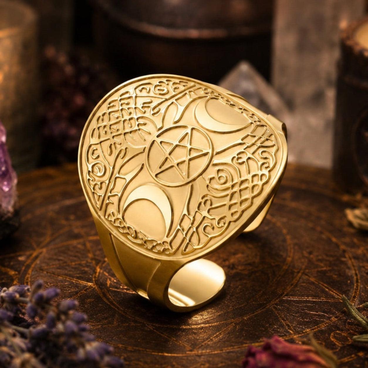 A gold-toned ring with intricate celestial and occult engravings, including a pentagram and crescent moon motifs, displayed on a dark wooden tray surrounded by candles, crystals, and dried botanicals.