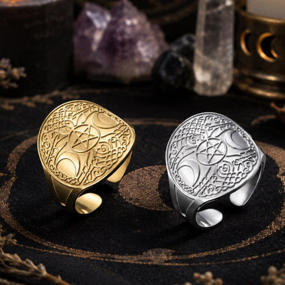 Two engraved occult-style rings, one gold and one silver, displayed on a dark patterned cloth with candles, crystals, and dried botanicals in the background.