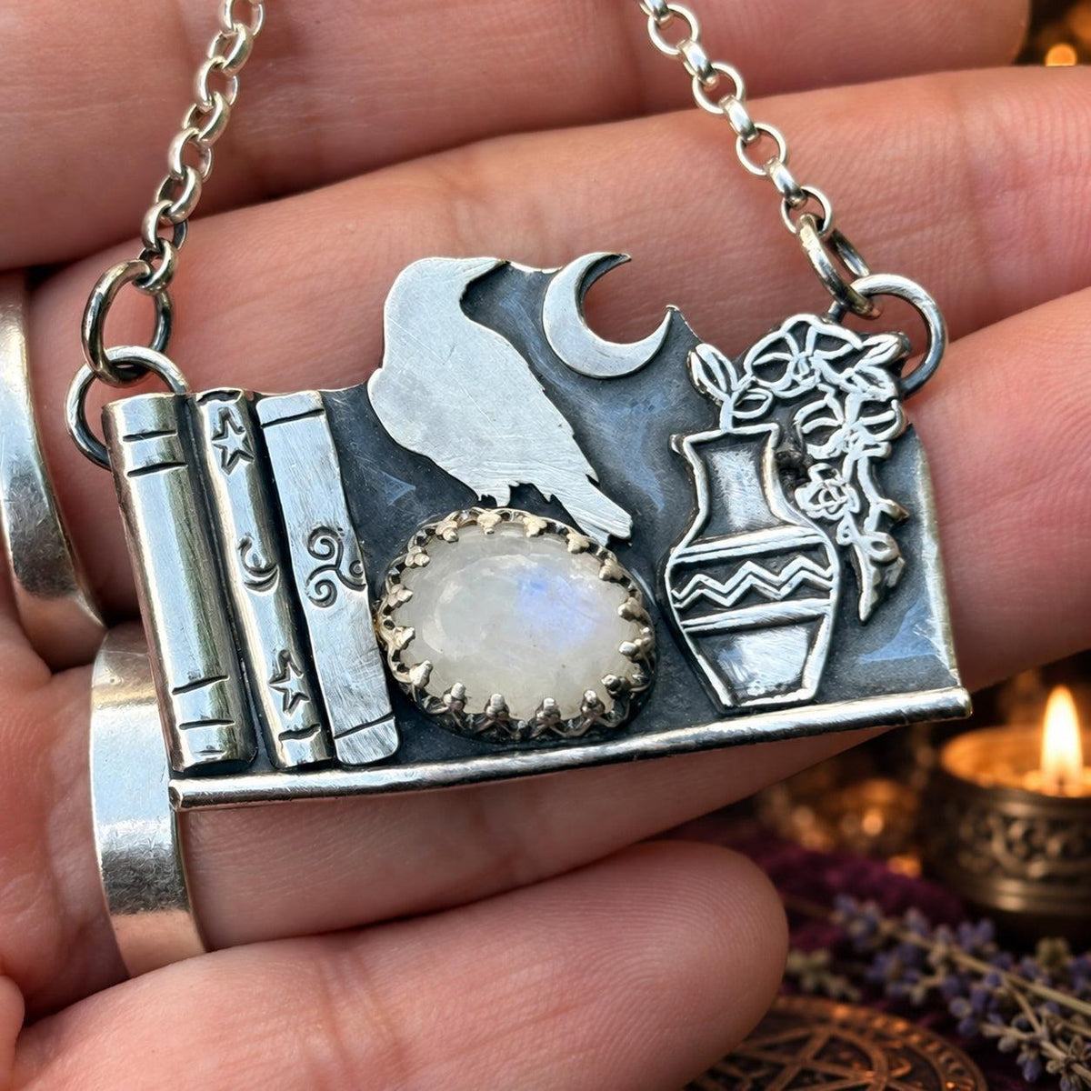 Raven bookshelf pendant necklace worn in hand with moonstone, crescent moon and vase - wiccan jewelry
