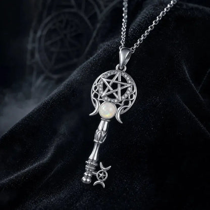 A silver-toned key-shaped pendant necklace with a pentagram design, moon phases, and a white opal-like center stone, displayed on a black velvet surface.