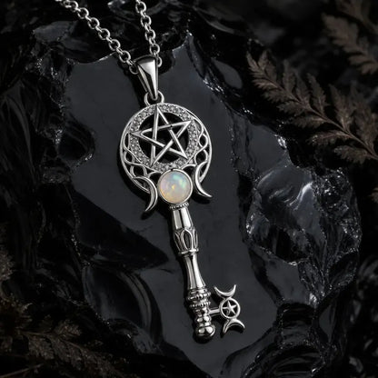 A silver-toned key pendant necklace with a pentagram and crescent moon design, featuring an opal inlay, displayed on a glossy black rock with dried botanicals.